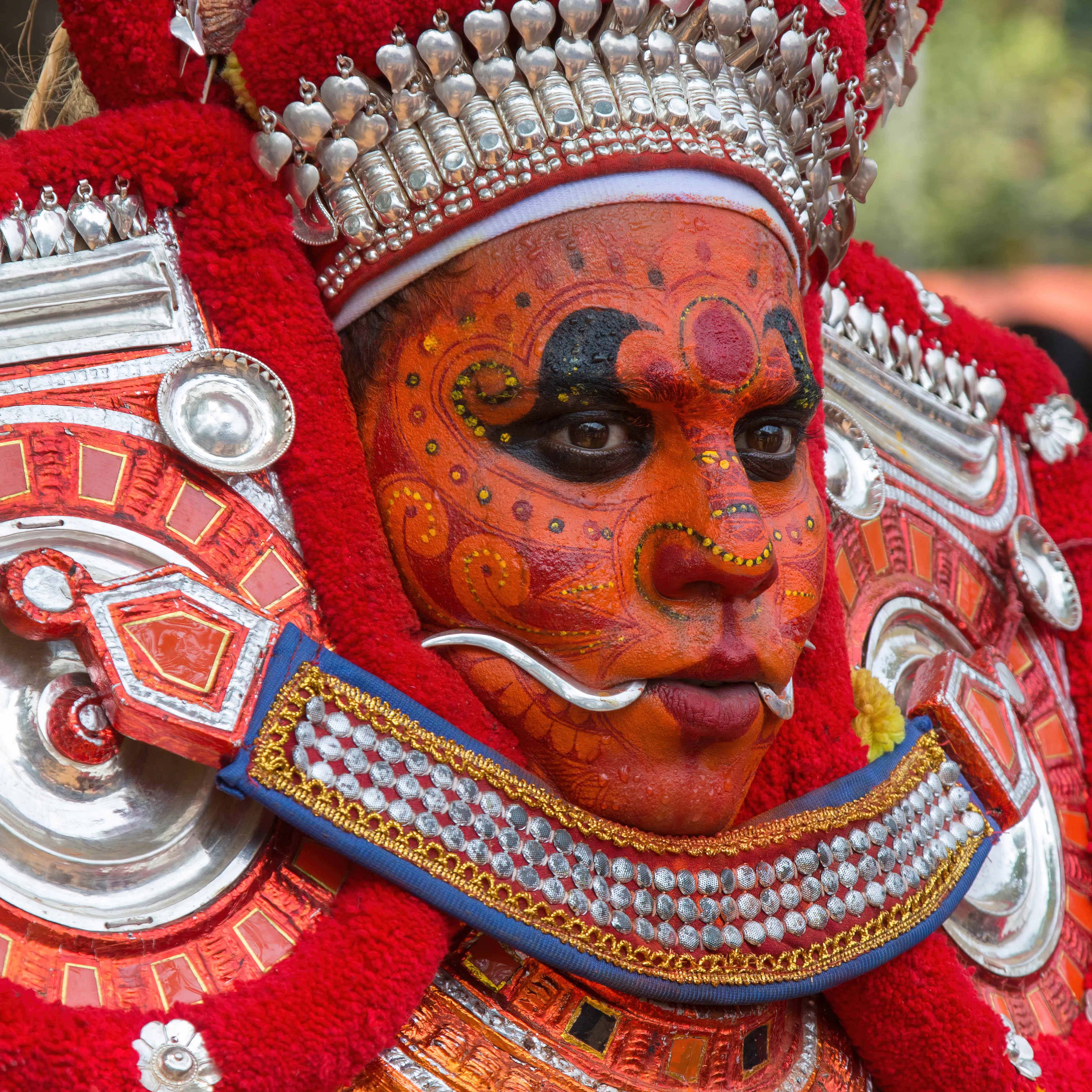 A Theyyam performer in red and orange ritual face paint and a silver headdress